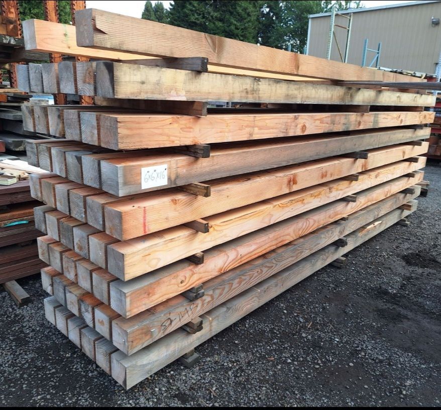 A large stack of long, rectangular wooden beams stored outdoors on a gravel lot