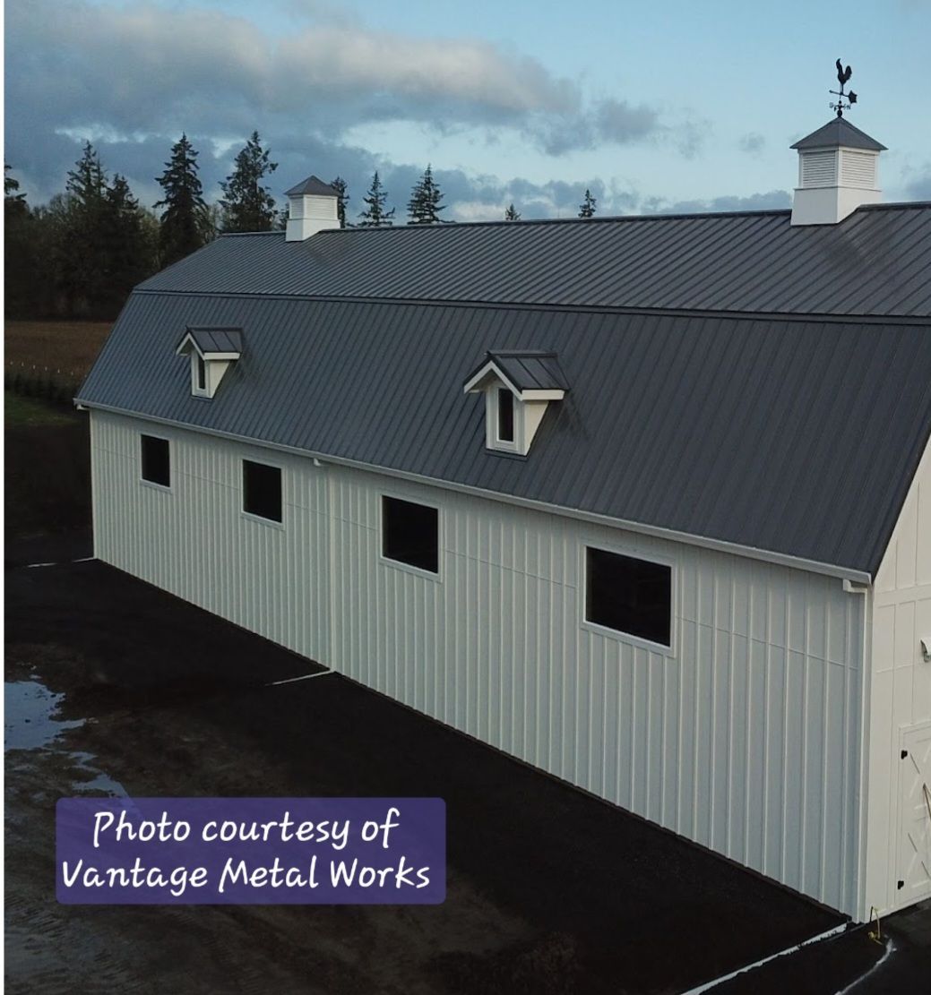 A white barn with a dark metal roof, cupolas, and small dormer windows under a cloudy sky.