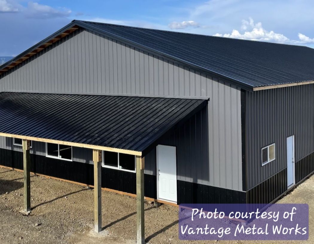 A grey and black metal warehouse with a dark roof and a porch supported by wooden beams under a blue sky.