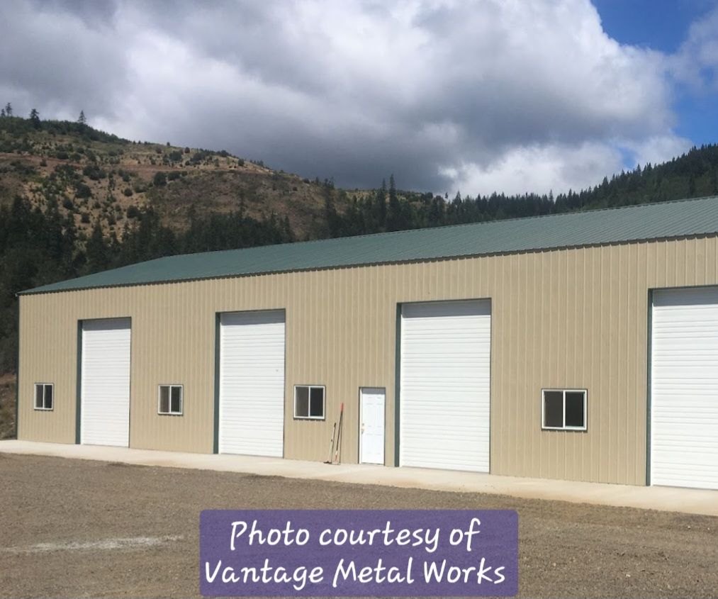 A long, tan metal warehouse with a green roof, four white roll-up garage doors, and several small windows against hills.
