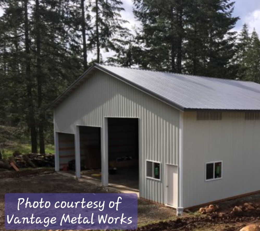 A white metal pole barn with a gray roof stands in a wooded area, featuring two large openings and a side entrance door.