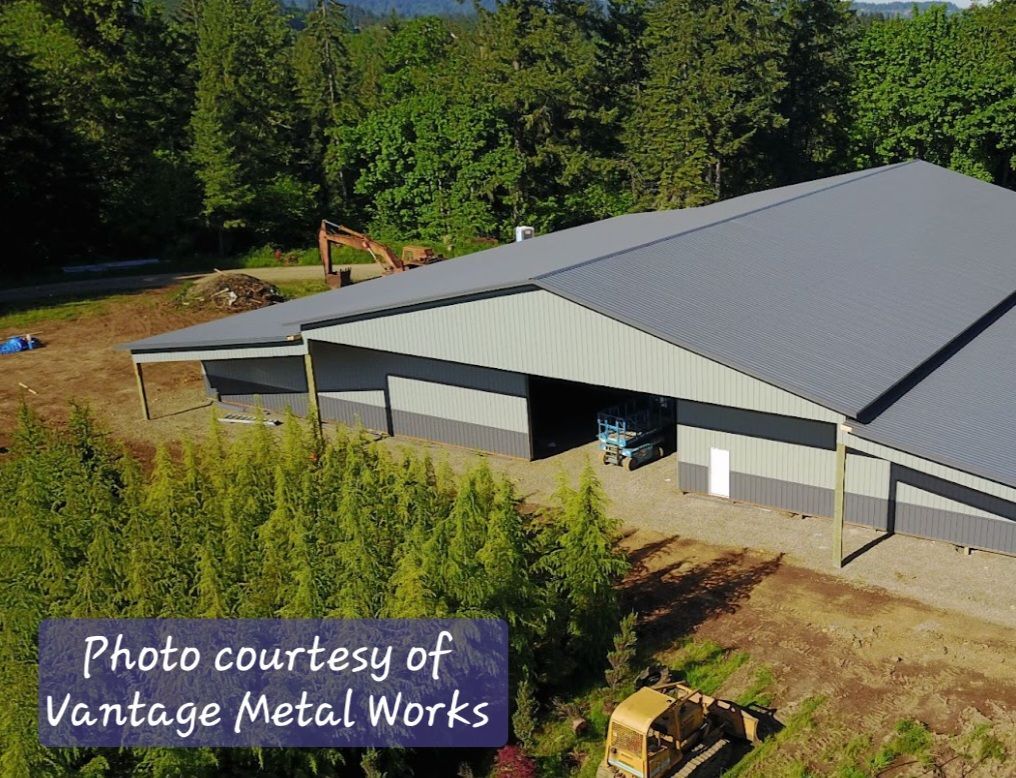 An aerial view of a gray metal building under construction near a forest, with construction equipment nearby.