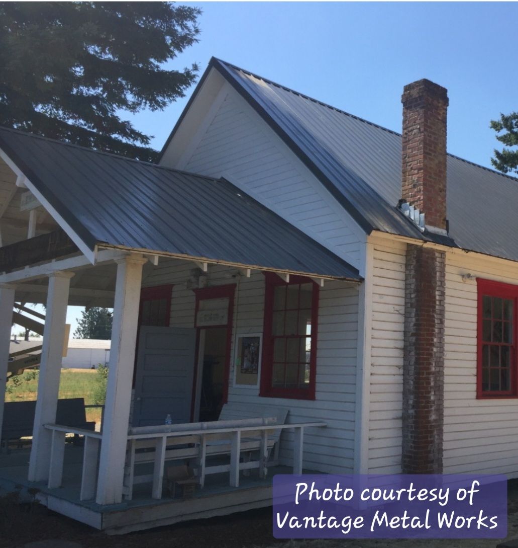 A white historic building with a dark metal roof, a front porch, and a brick chimney under a clear blue sky.