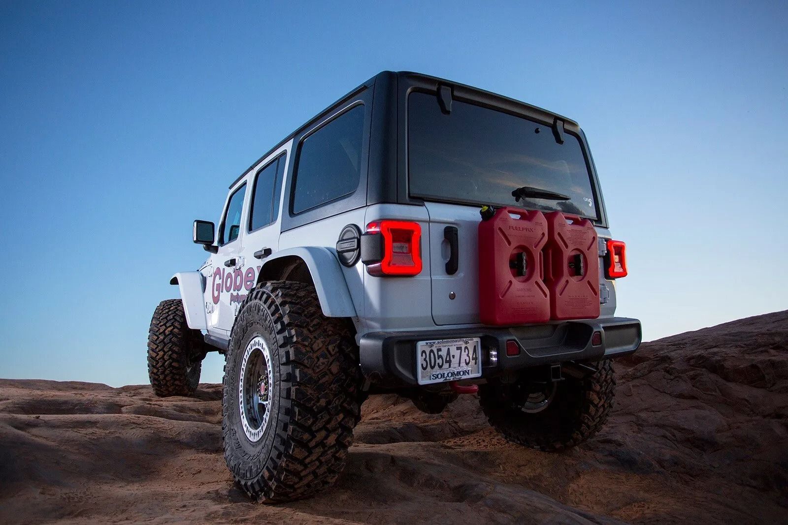 White Jeep Wrangler climbing a rocky hill with two red gas cans attached to the rear.