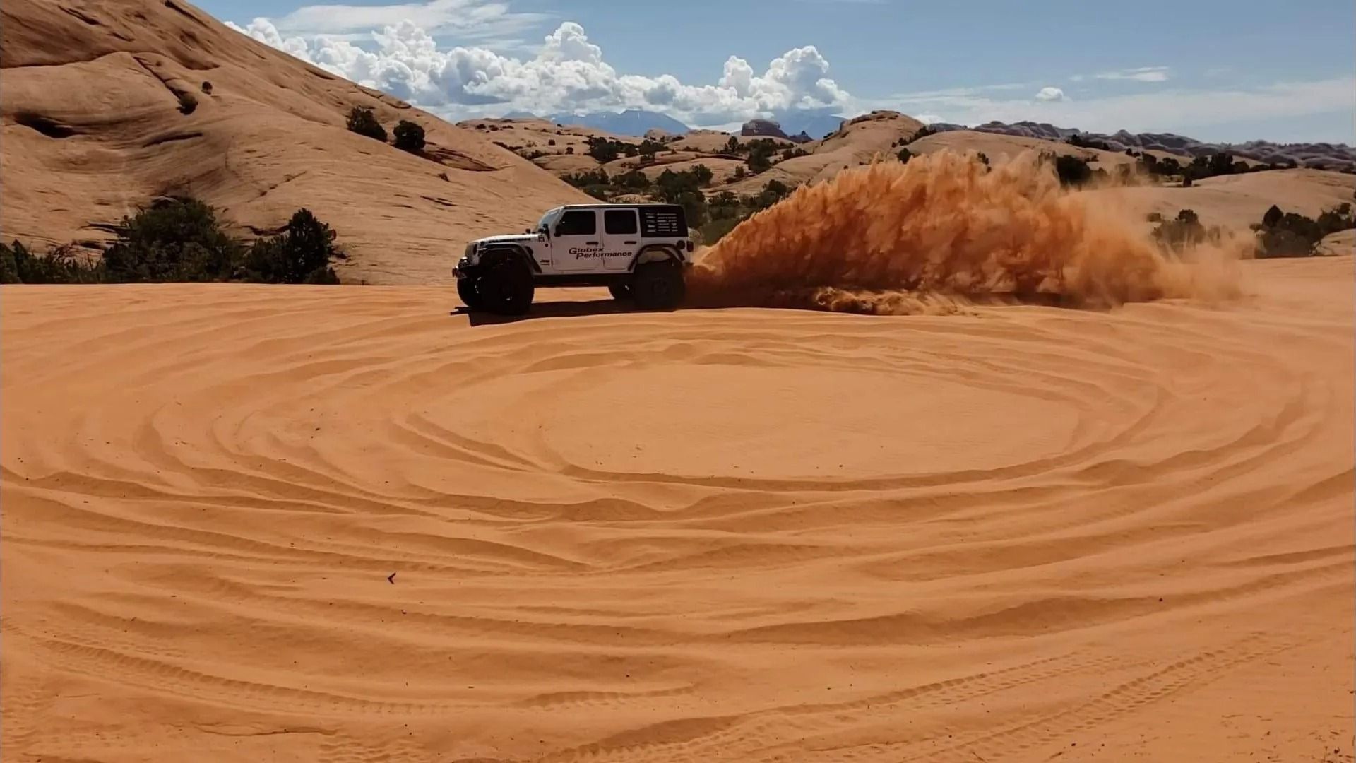 White SUV drifts in a circle on red sand, kicking up a large cloud of dust.