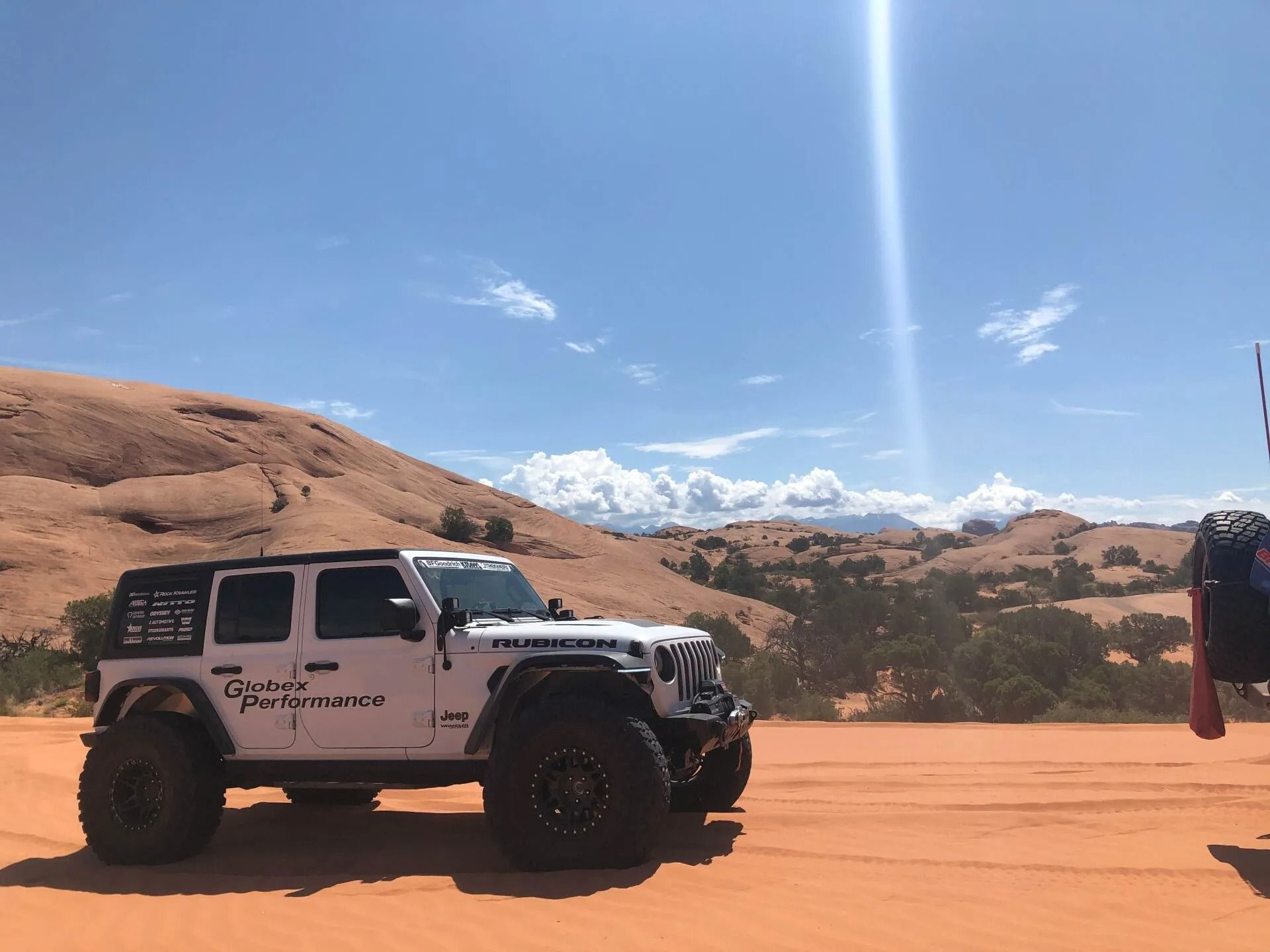 White Jeep Wrangler with large tires on red sand, sunny day in desert landscape.