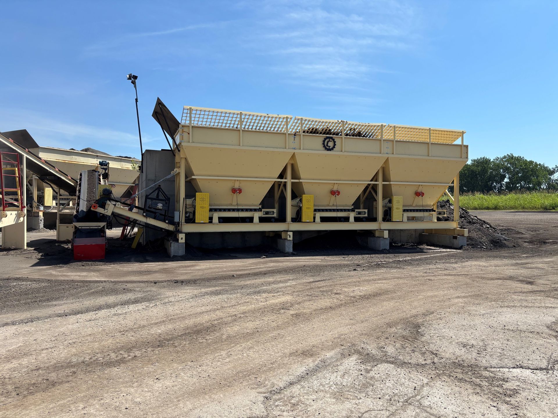 Yellow industrial machine on gravel lot under a blue sky, with a conveyor and covered bins.