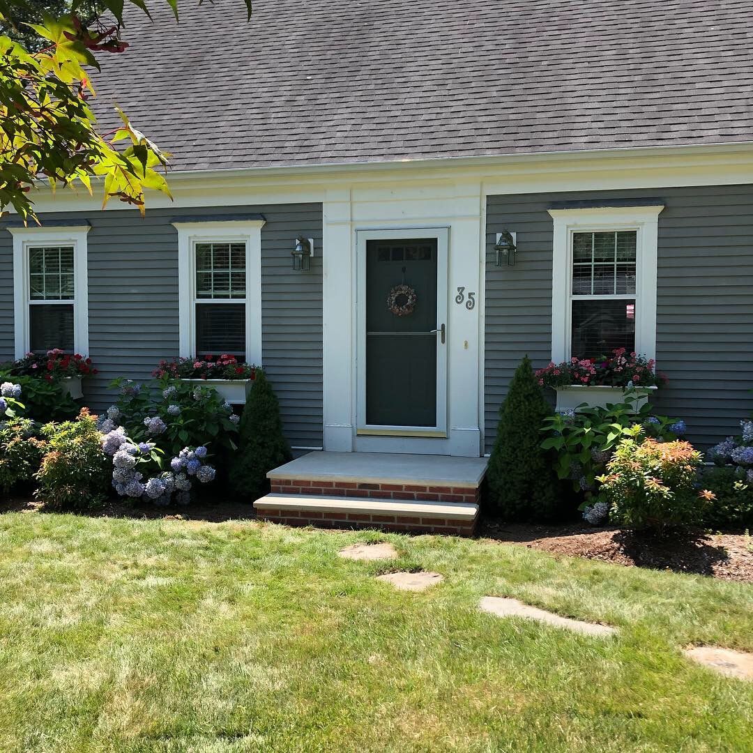A gray house with a white door and windows is surrounded by flowers and bushes