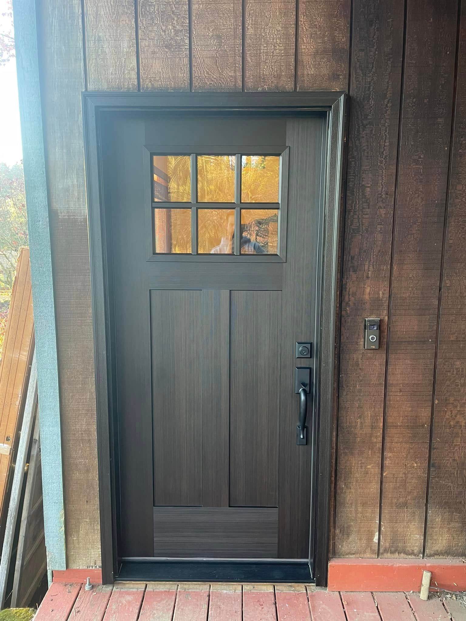 Dark brown door with a square window at the top, surrounded by brown trim, set in a wooden wall. A black handle and lock are visible.