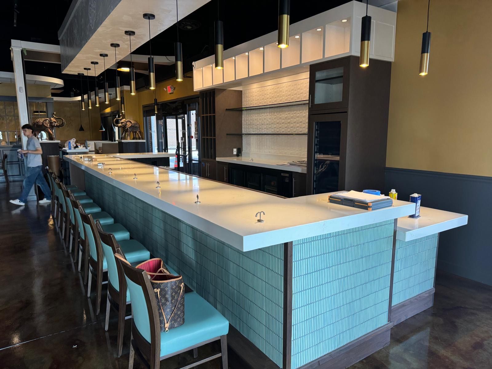 Modern bar interior with a long teal counter, hanging lights, and shelves behind the bar