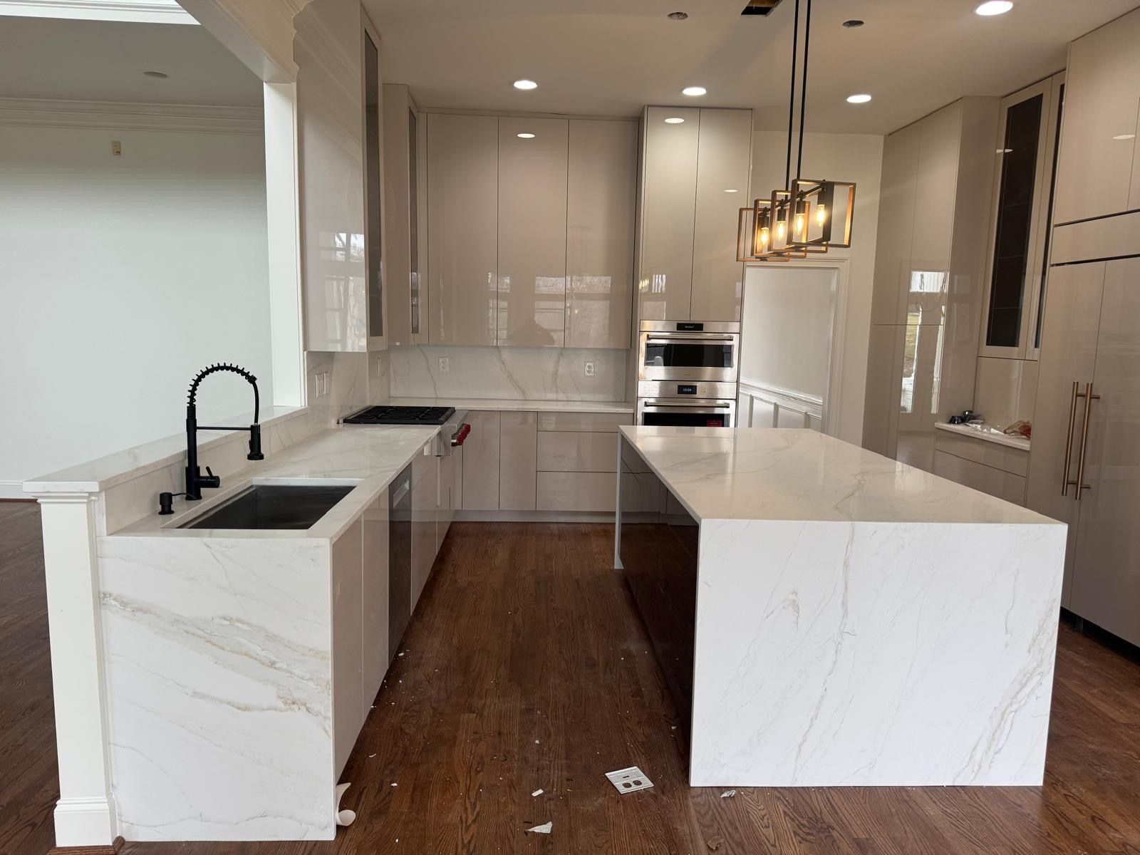 Modern white kitchen with marble island, dark wood floor, built-in oven, and black sink faucet.