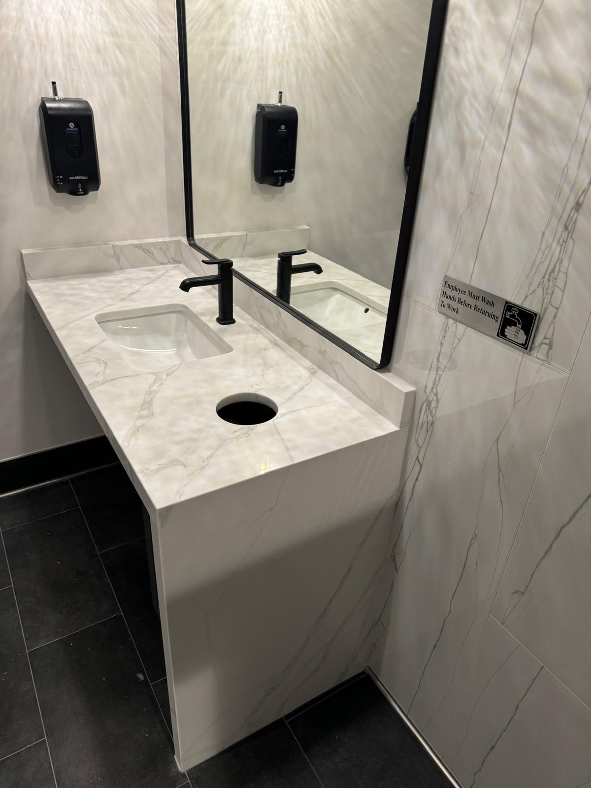 Modern restroom sink with white countertop, black faucets, mirrors, and wall-mounted soap dispensers