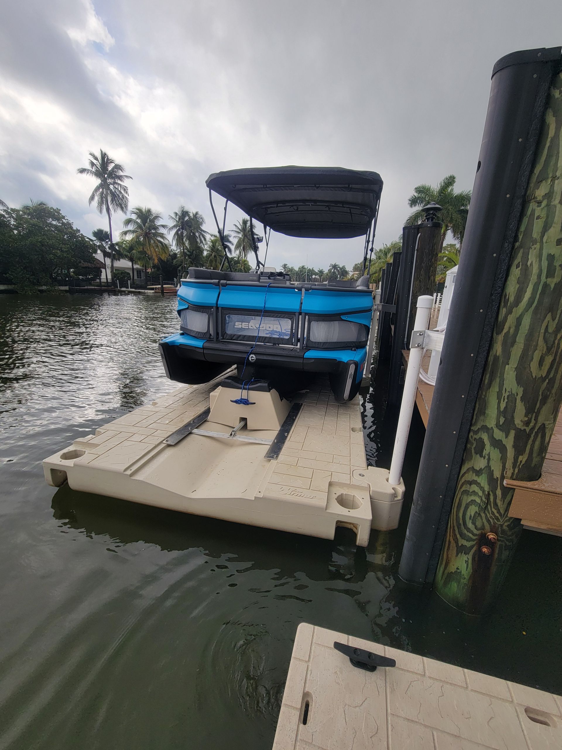 Blue pontoon boat on a beige floating dock, near a wooden dock with a camouflaged support. Overcast sky.