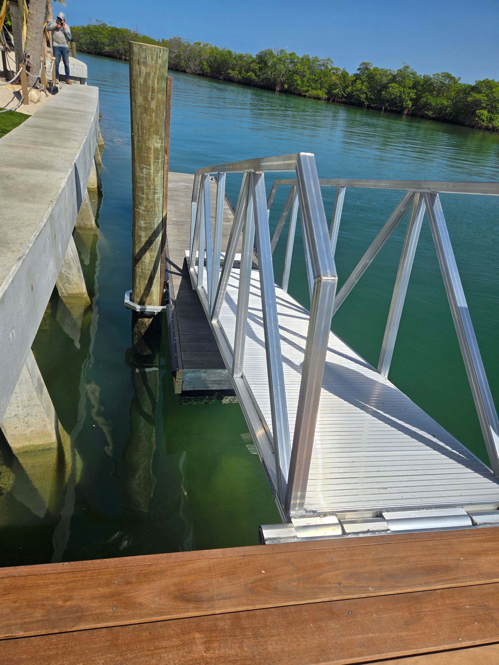 Aluminum ramp leading to a dock over green water, with a wooden dock and mangrove trees in the background.