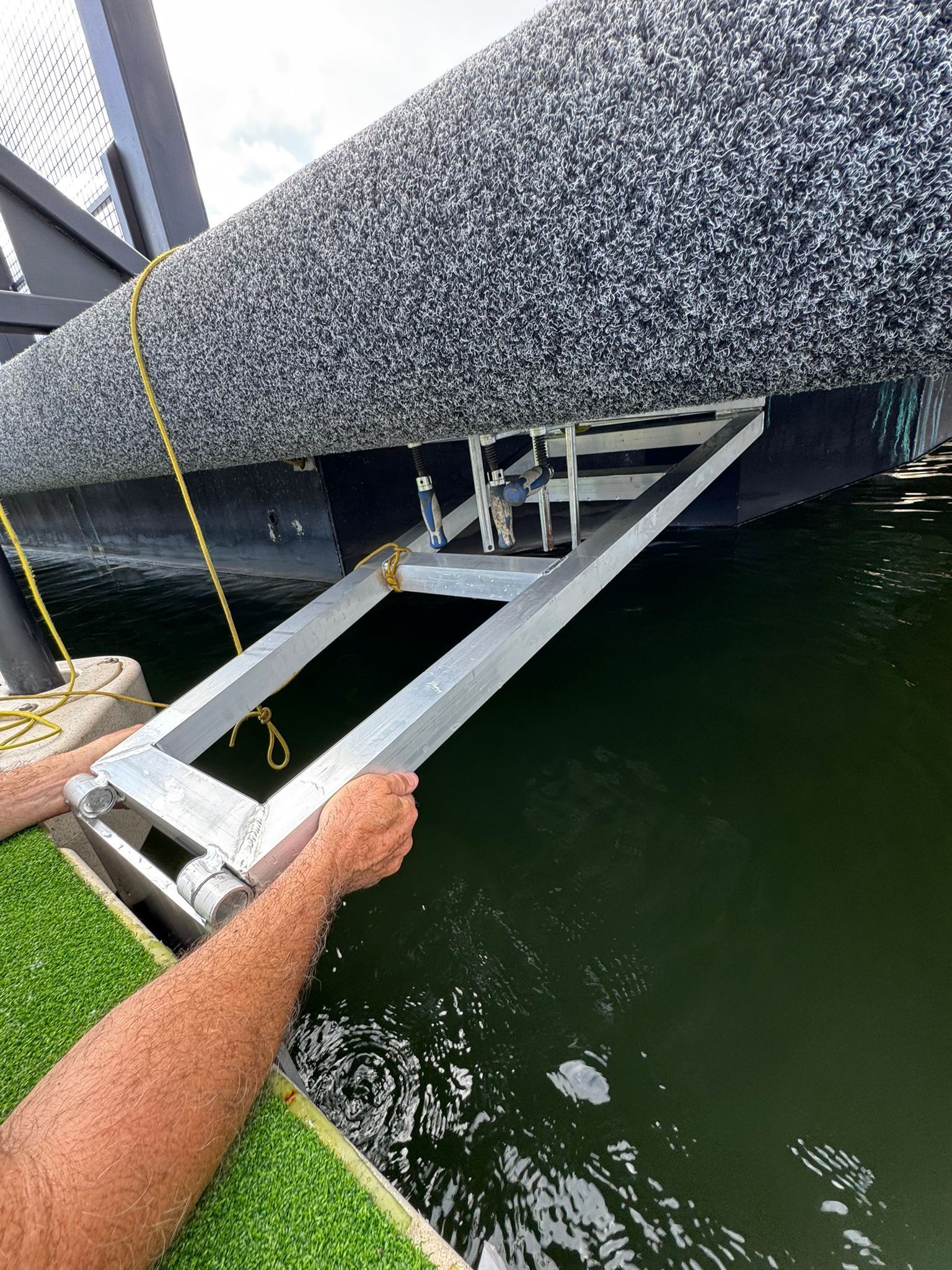 Person adjusting a ramp attached to a black and white pontoon boat, over green water.