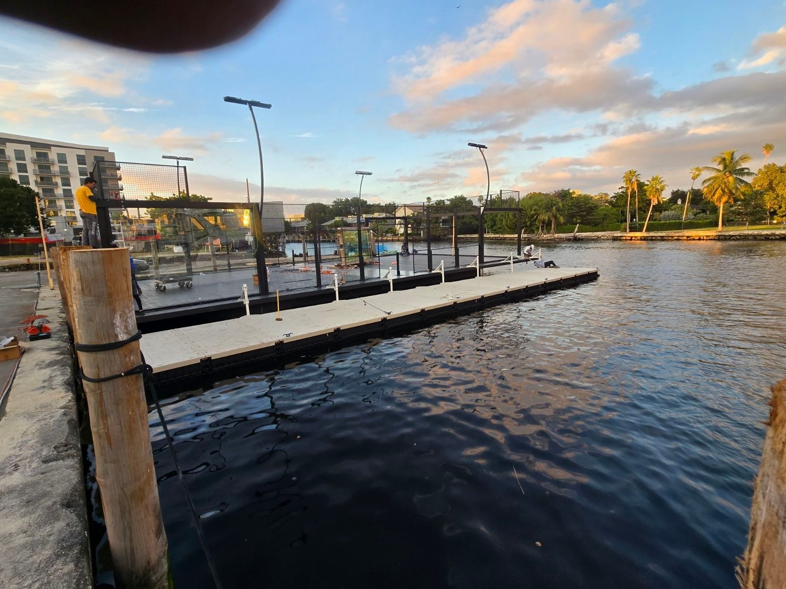 Boat docks on a waterway under a cloudy sky. Palm trees and buildings are visible in the background.