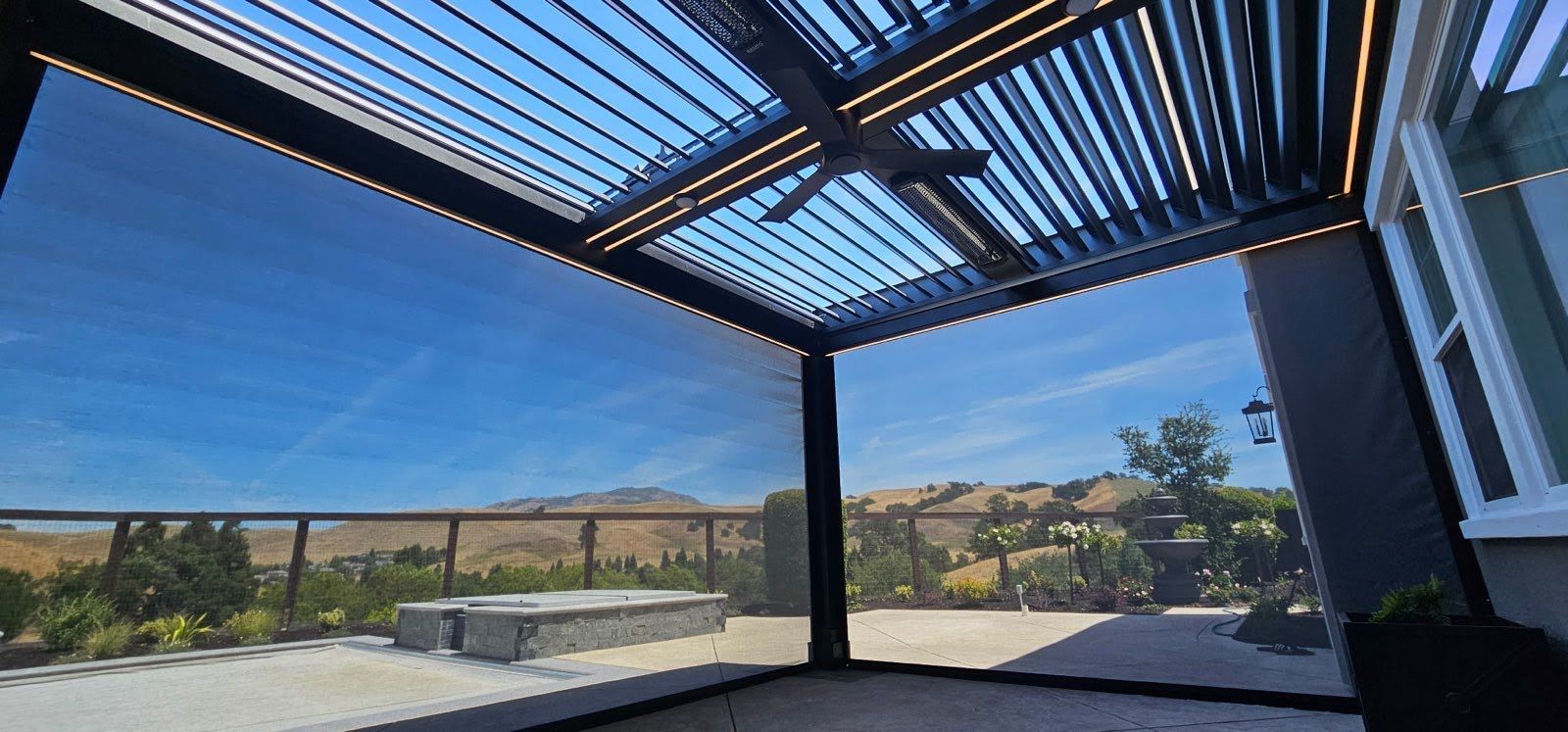 Covered patio overlooking a sunny backyard and hills, with a pergola roof and black railings