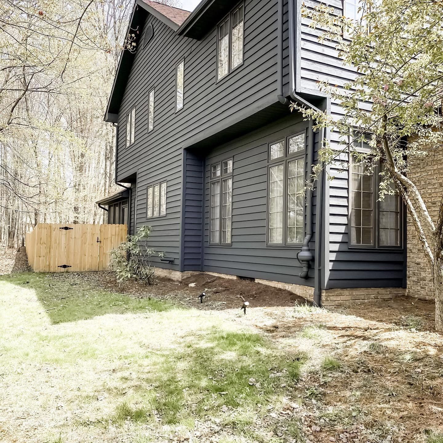 Dark gray two-story house with white trim, a small yard, and a wooden fence beside trees