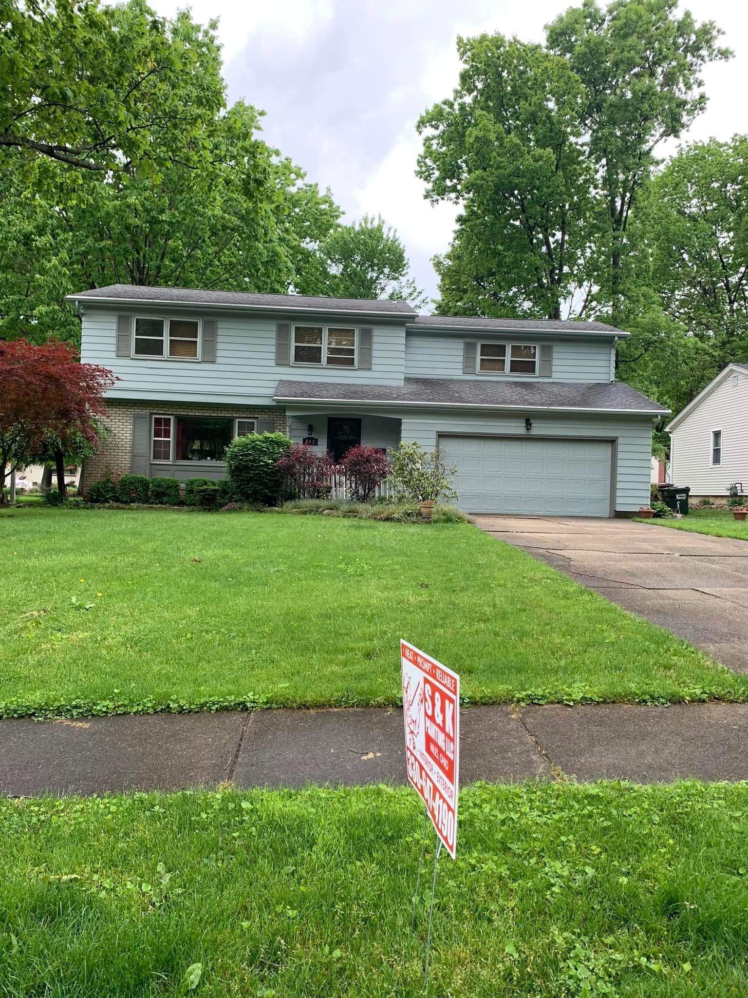Two-story gray house with attached garage, green lawn, and a red yard sign in front.