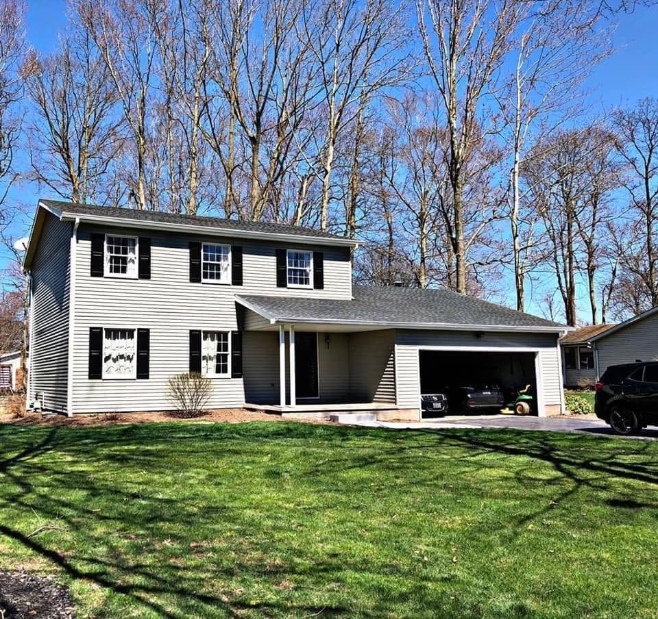 Two-story white house with black shutters, attached carport, and green lawn under a clear blue sky