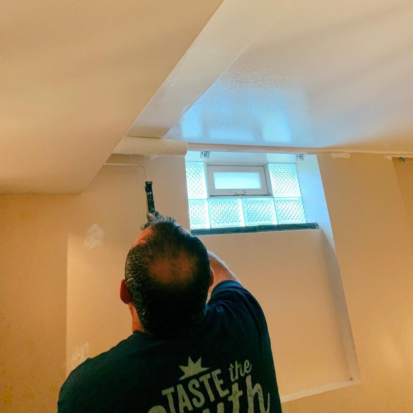 Person using a paint roller to coat a white ceiling near a small window.