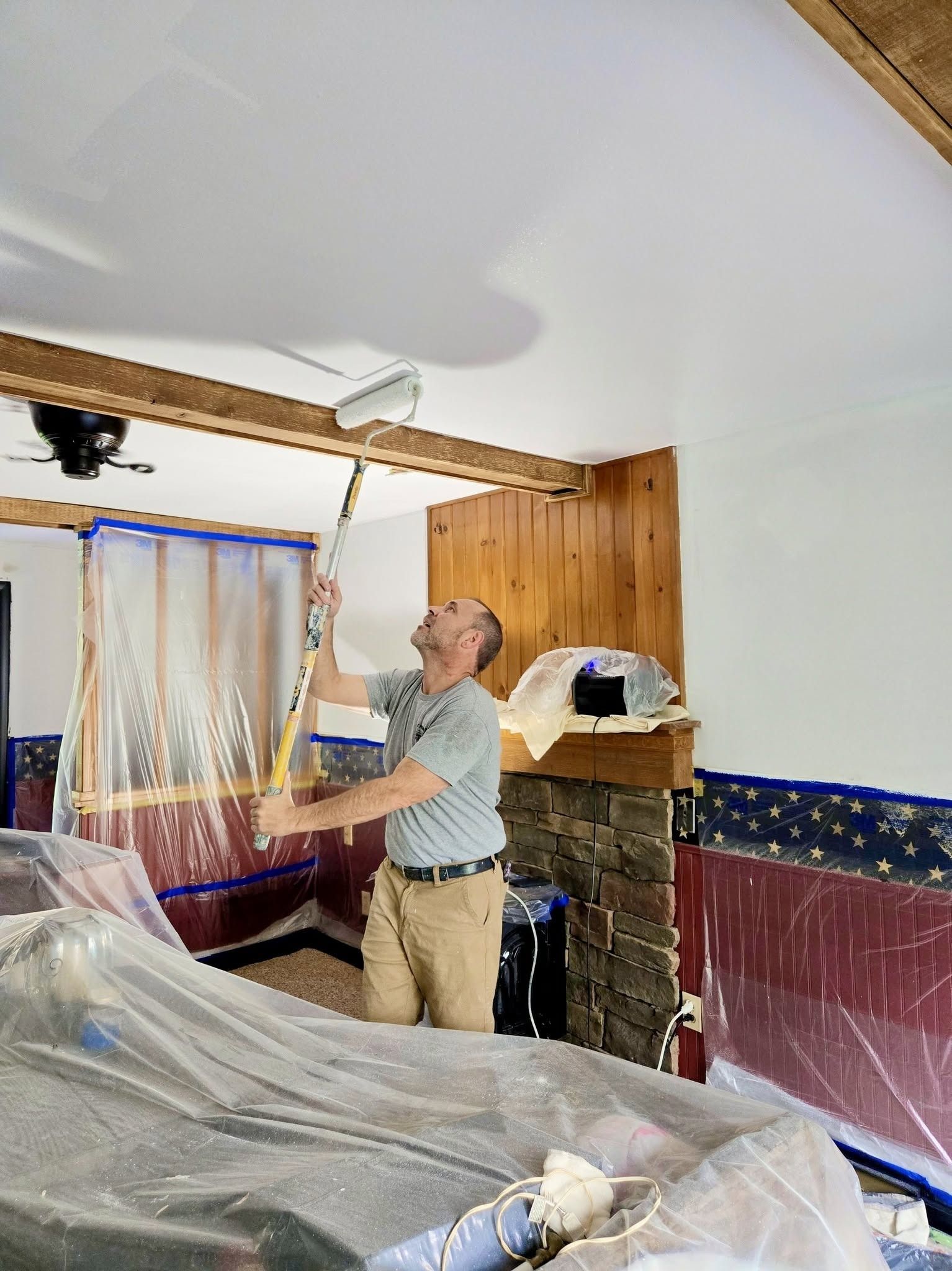 Person in a covered room painting a ceiling near a window and brick fireplace.
