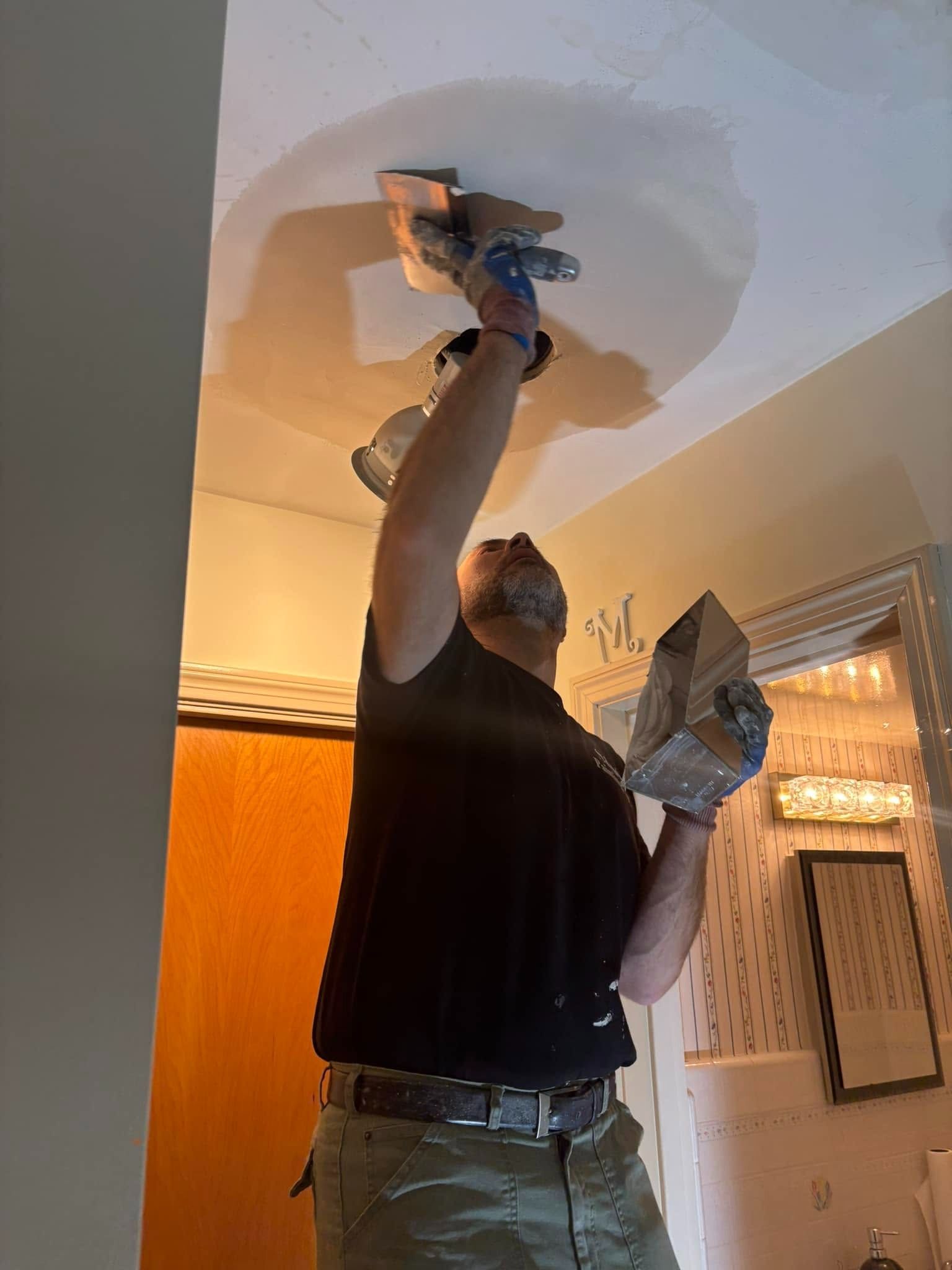 Person plastering a ceiling hole with a putty knife, holding a repair product in a living room.