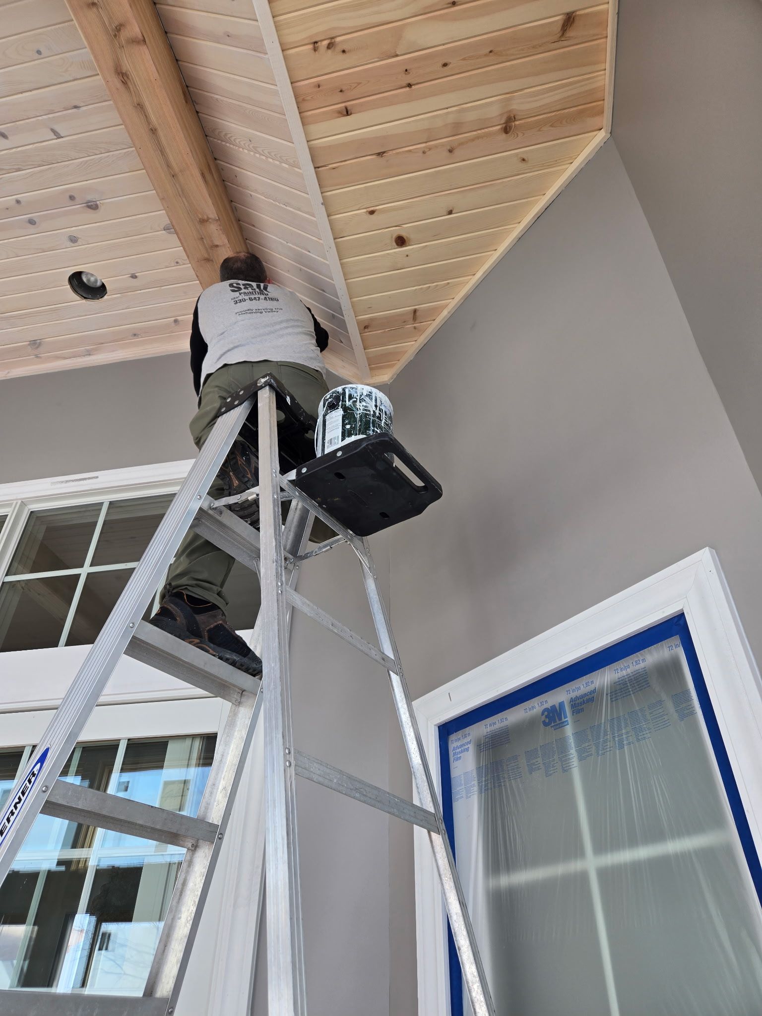 Worker on a ladder painting a wooden porch ceiling near a window