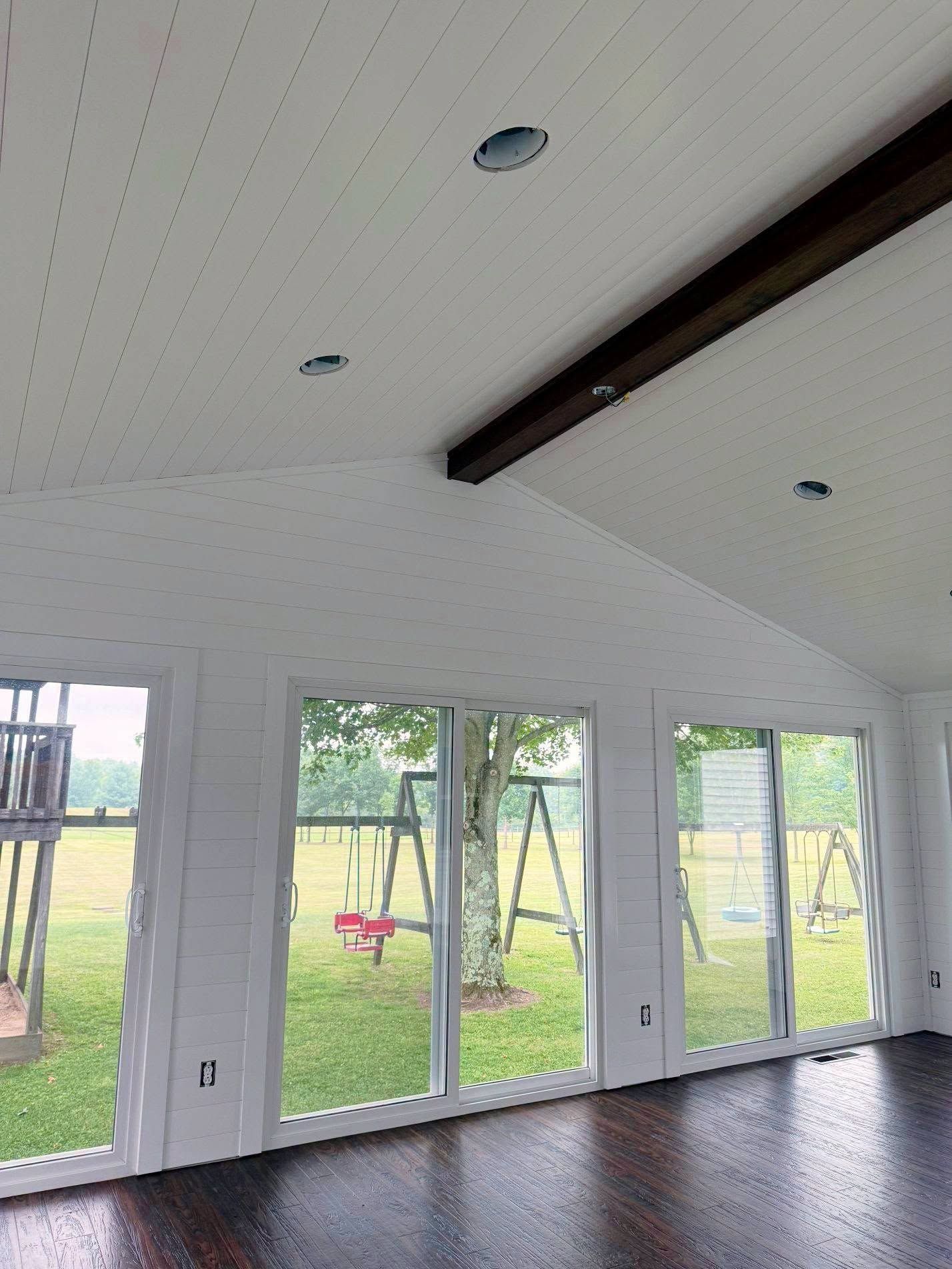 Bright, empty room with vaulted white ceiling, dark floor, and glass doors overlooking a grassy yard.