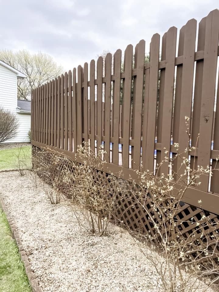 Brown wooden fence along a gravel path beside a bare hedge and grassy yard.