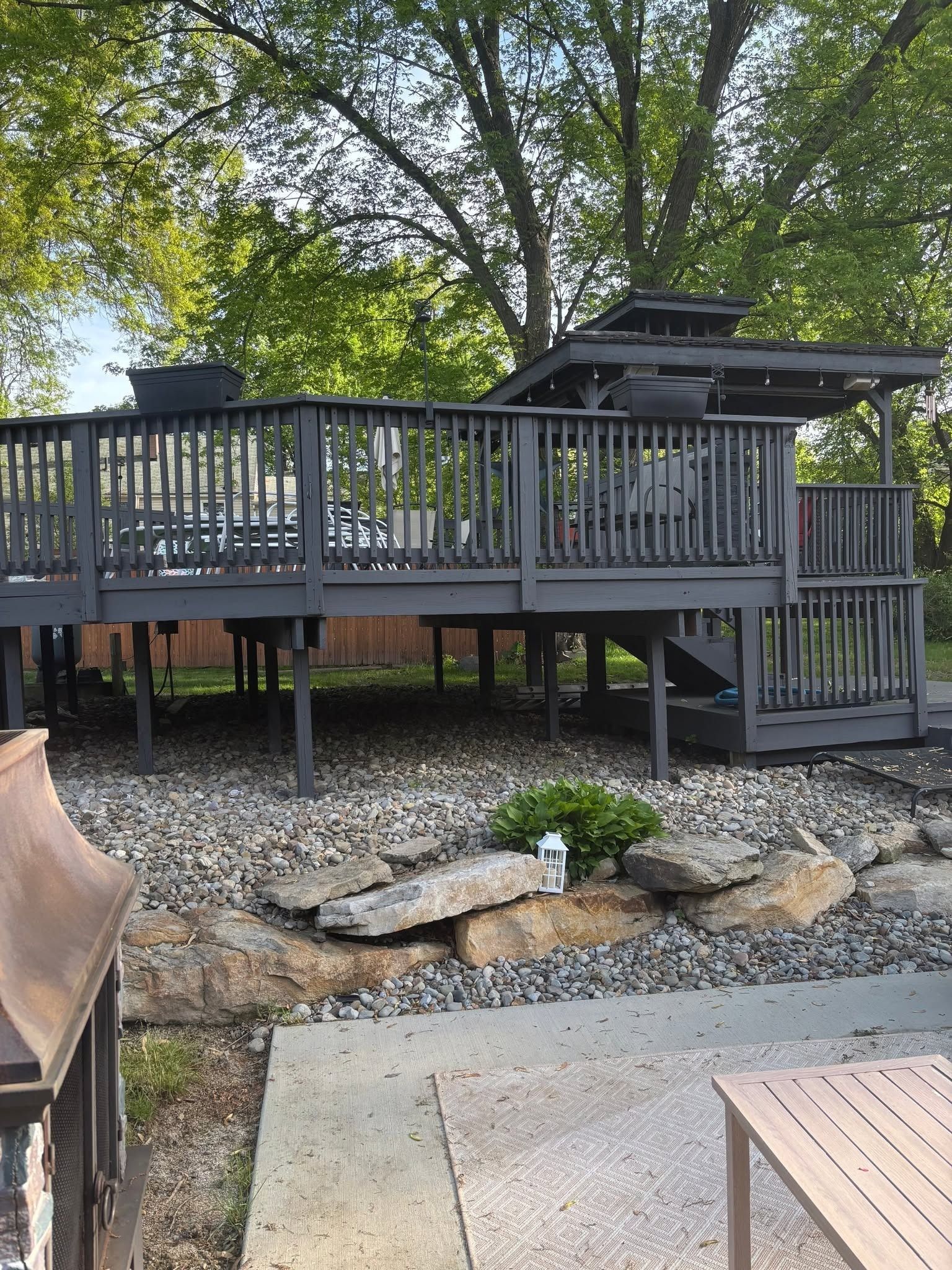 Backyard deck with gray railing and stairs above a stone garden and patio, surrounded by trees