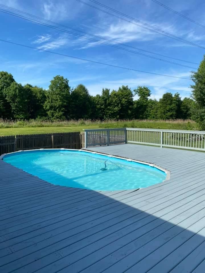 Backyard deck with an empty oval swimming pool and green trees under a blue sky