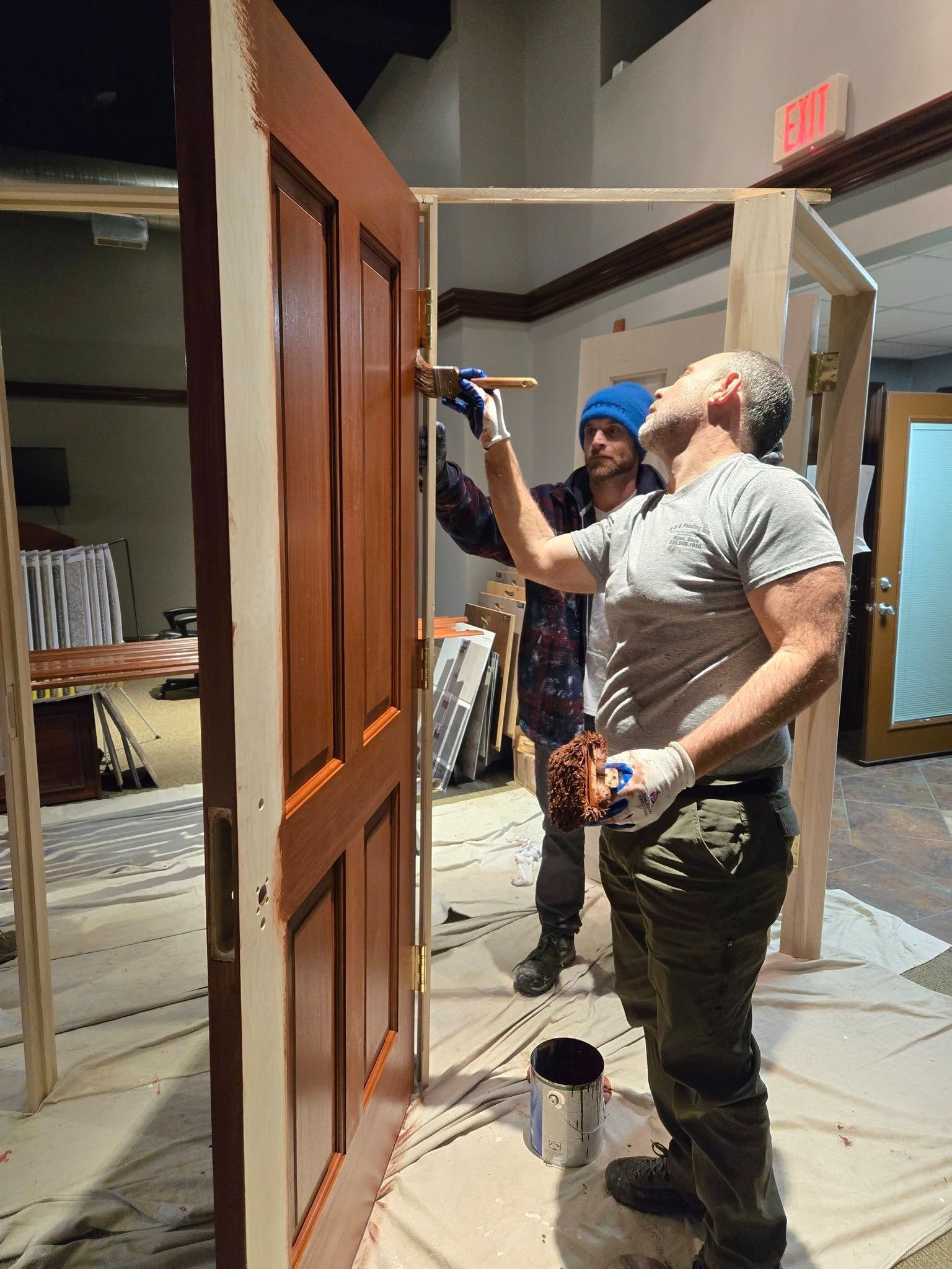 Two men painting a wooden door frame indoors, with paint cans and tools nearby.