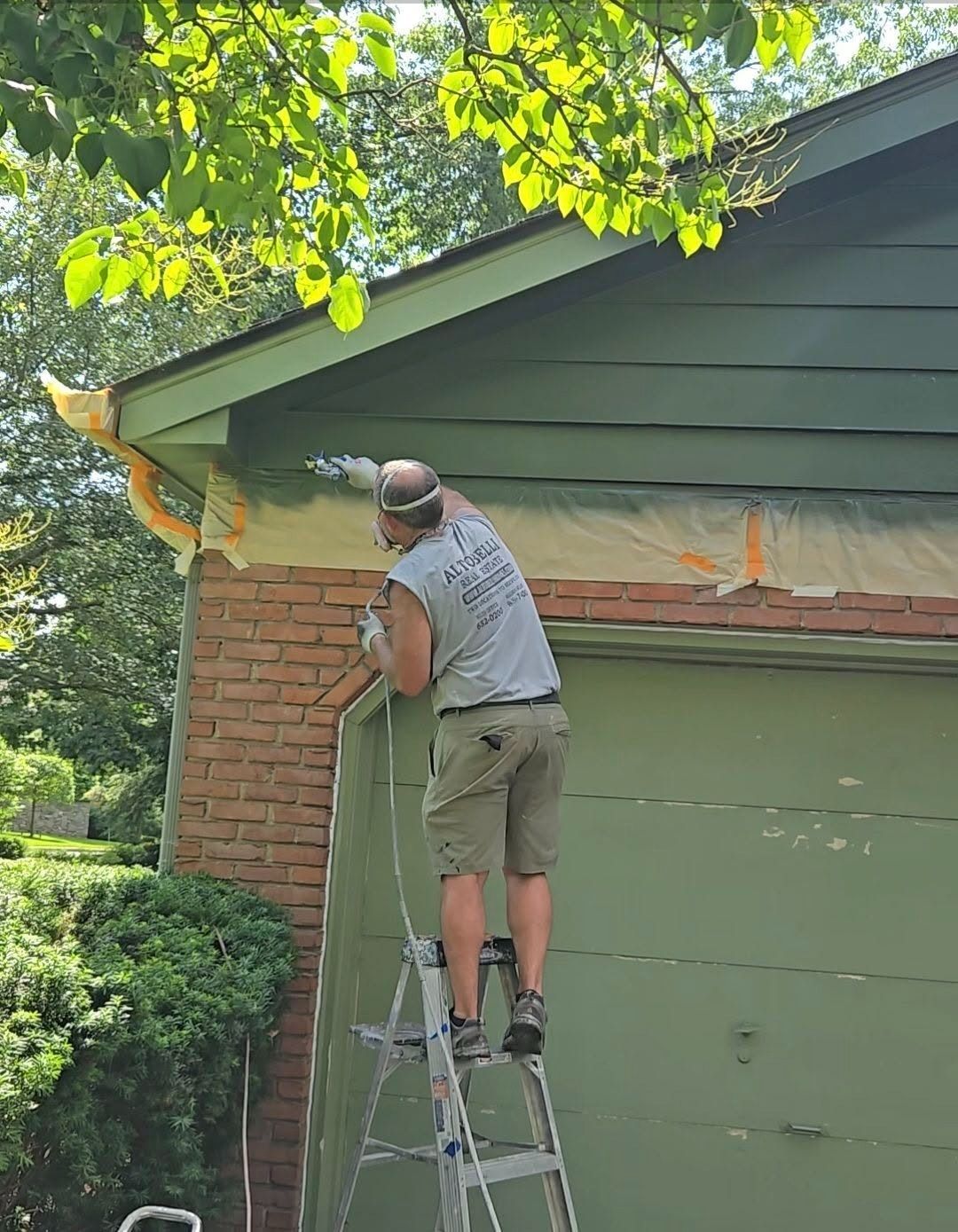 Person on a ladder painting the trim of a house near a green garage and brick wall