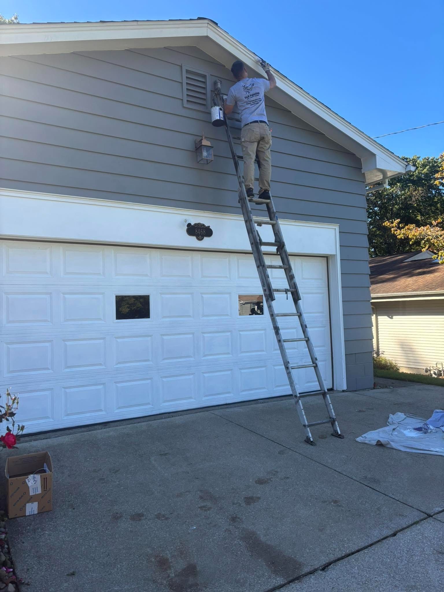 Person on a ladder painting the upper exterior wall of a house above a garage.