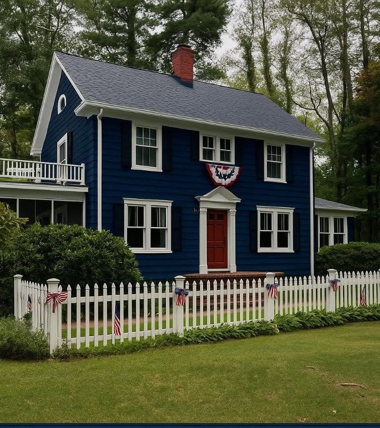 Blue colonial-style house with white trim, red door, white picket fence, and patriotic bunting by trees