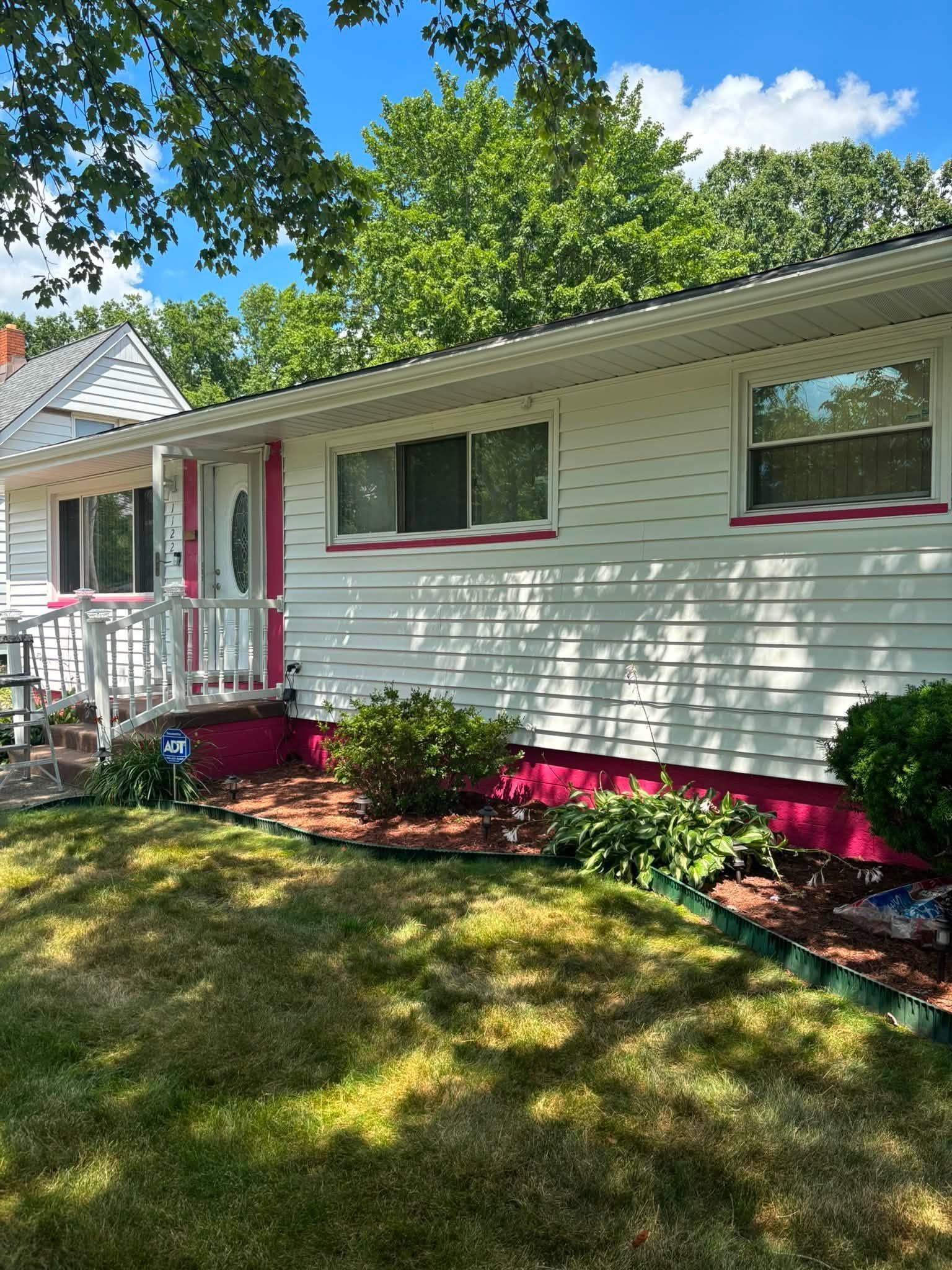 Single-story white house with red trim, front porch, and landscaped flower beds in bright sunlight