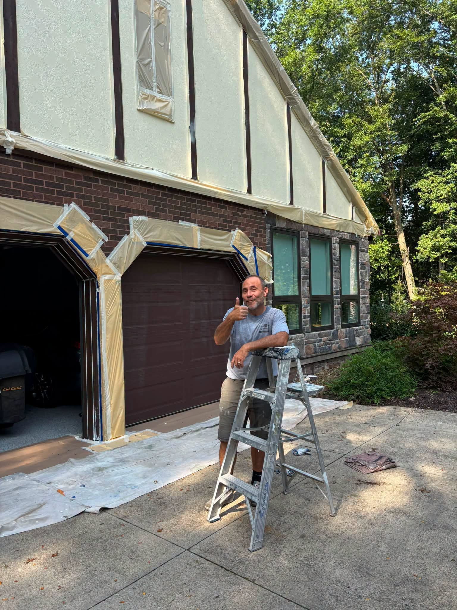 Person standing on a ladder beside a house under renovation, painting exterior trim.