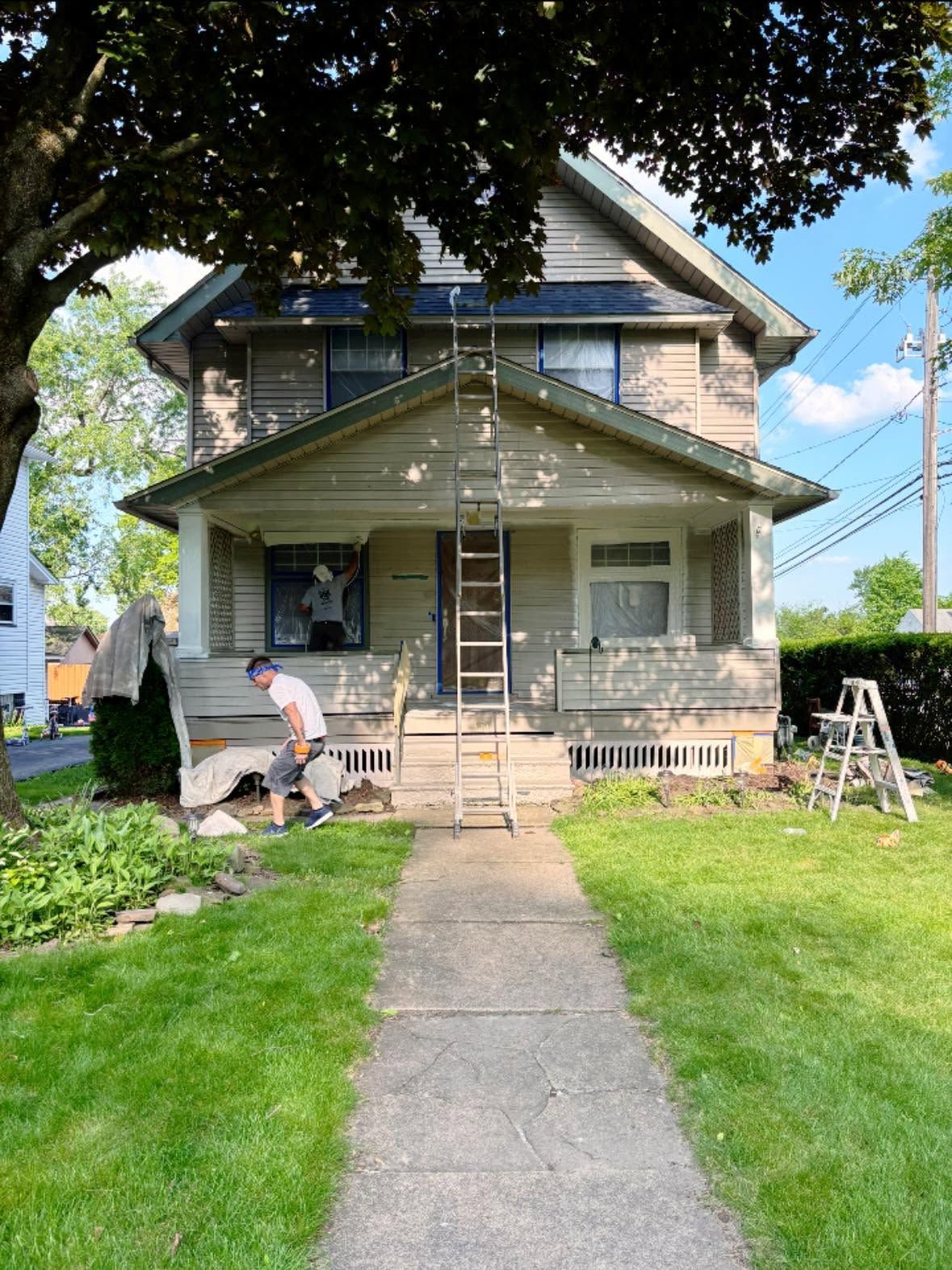 Workers painting a two-story house with ladders and supplies in the front yard