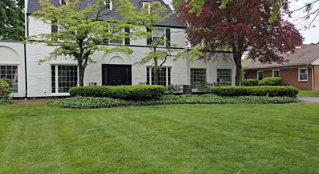 White house with black shutters behind a green lawn and hedges under trees