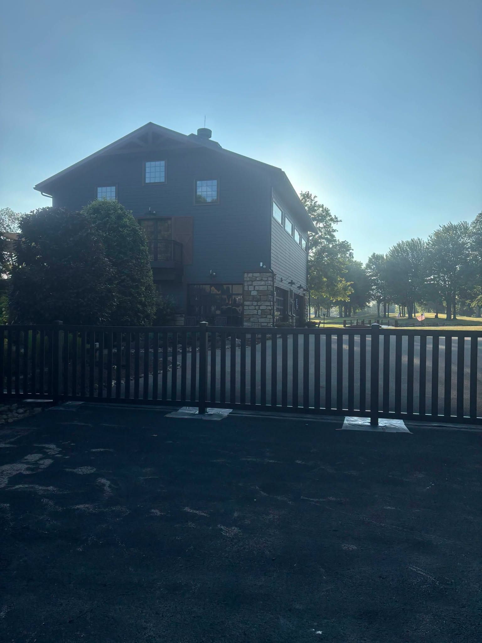 A house behind a dark fence in bright sunlight, with trees and open field in the background.