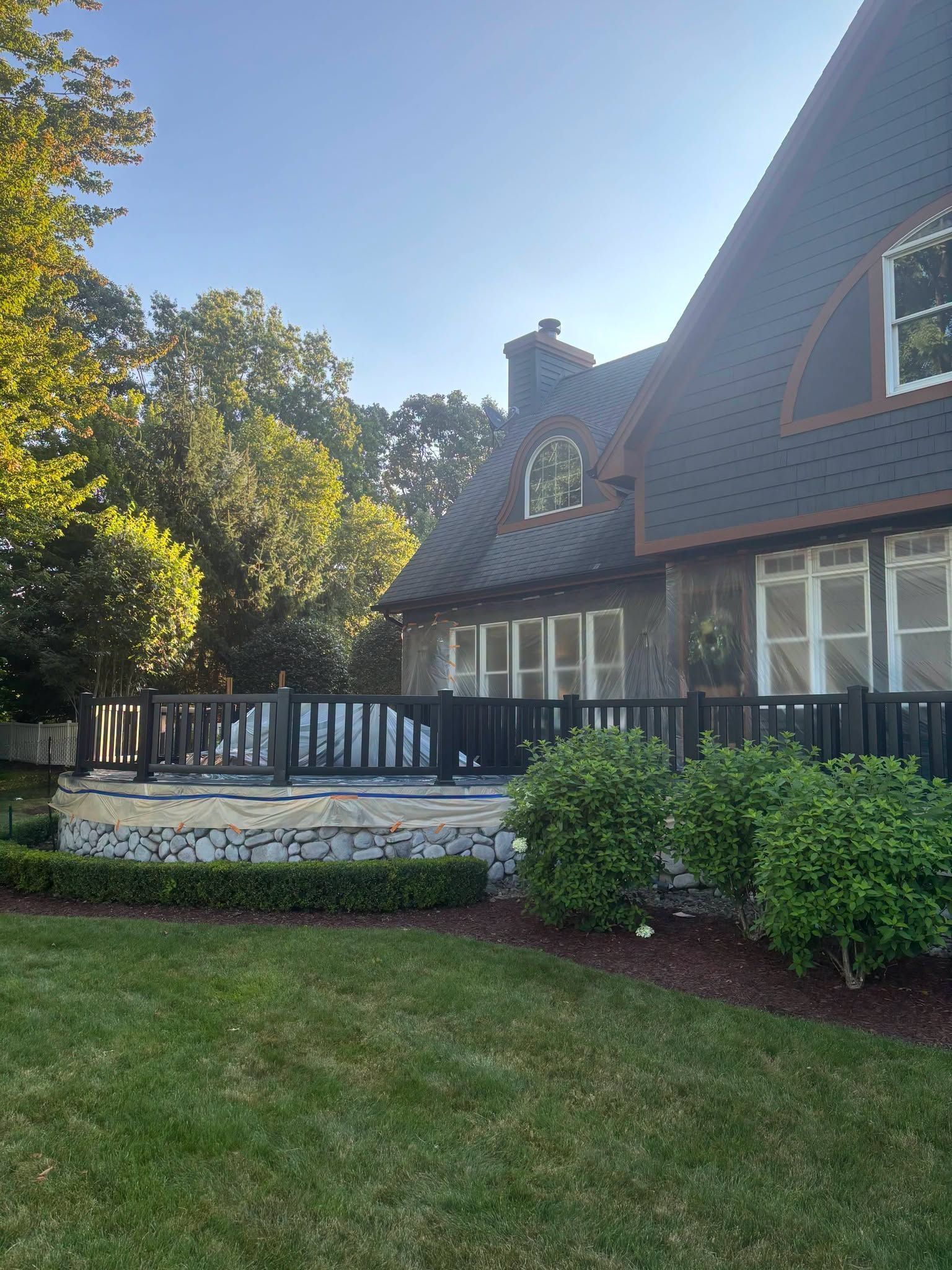 A house with dark roof, front deck, and green lawn beside trees in sunlight