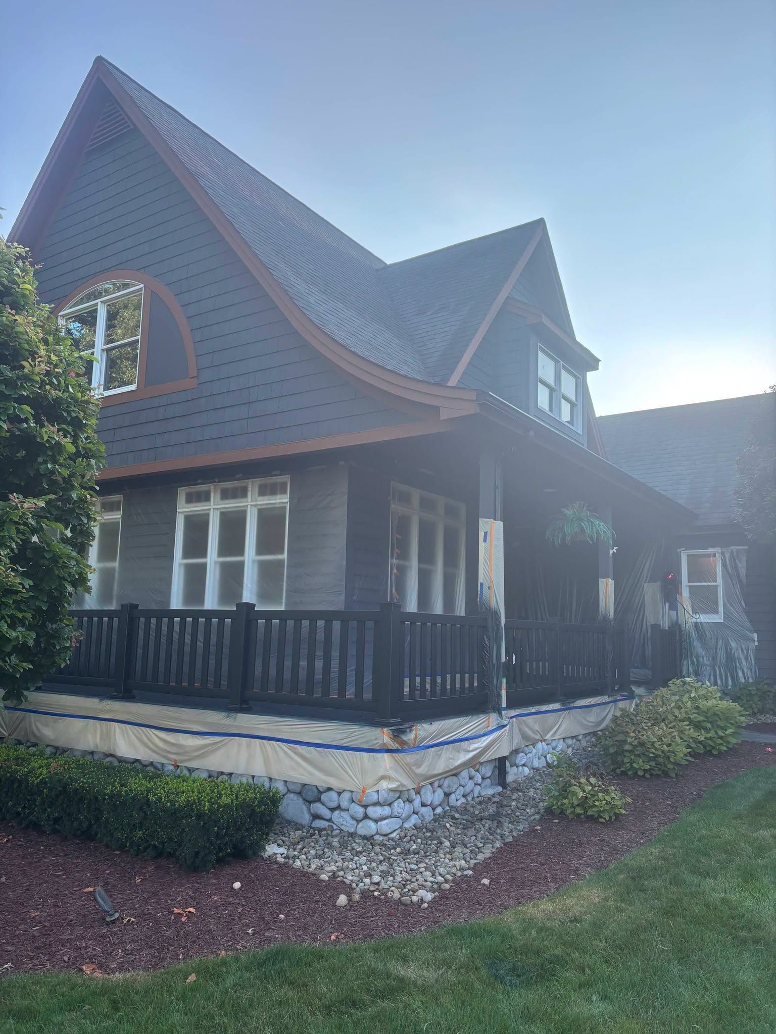 A house with steep roof, stone foundation, and black porch railings in a landscaped yard