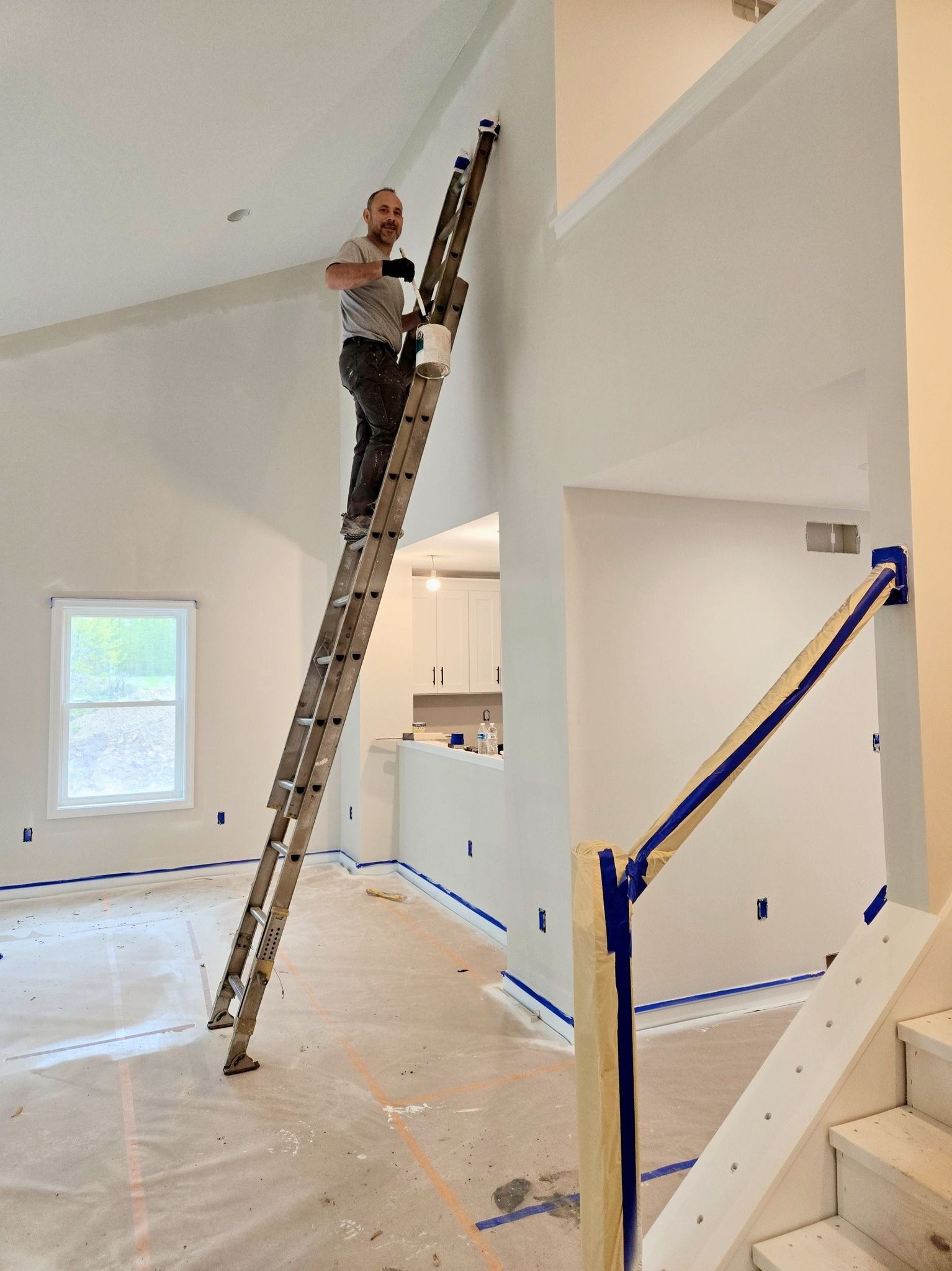 Worker on a tall ladder painting a bright interior room with blue painter’s tape on the walls.