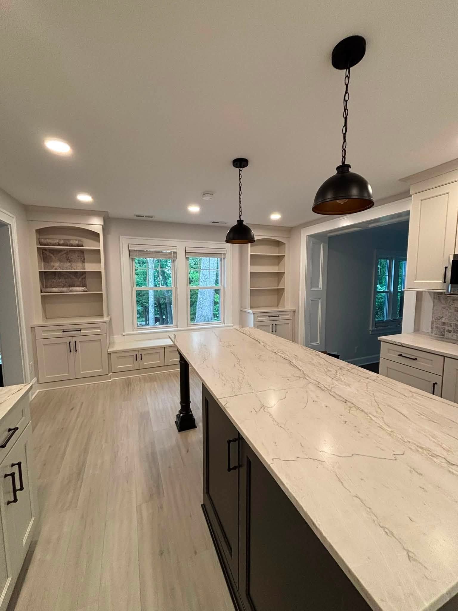 Bright white kitchen with marble island, black pendant lights, built-in shelving, and large windows