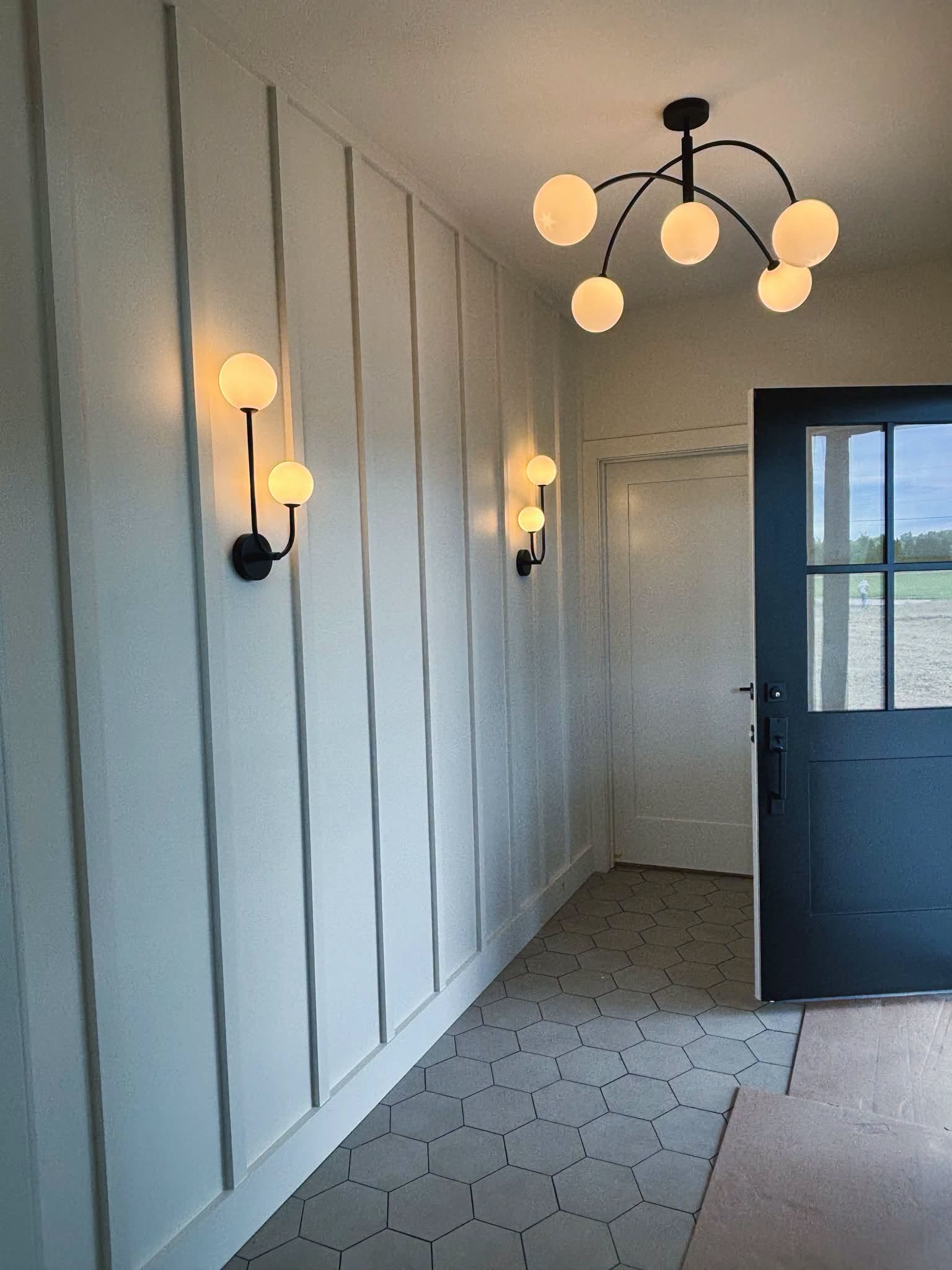 Bright white hallway with wall sconces, black-and-gold ceiling light, patterned tile floor, and an open blue door.