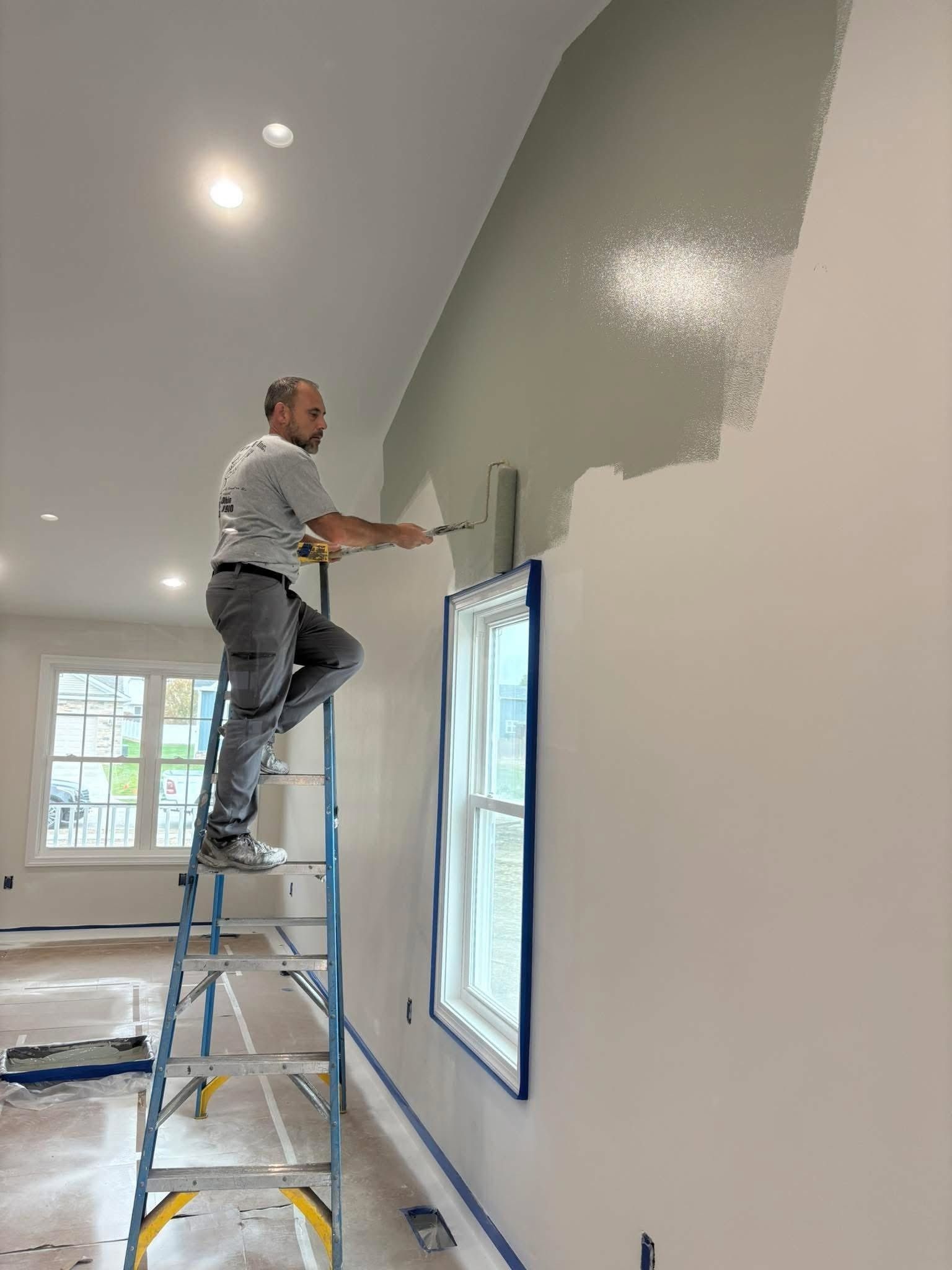 Person on a ladder painting a high wall white in a bright room with a window.