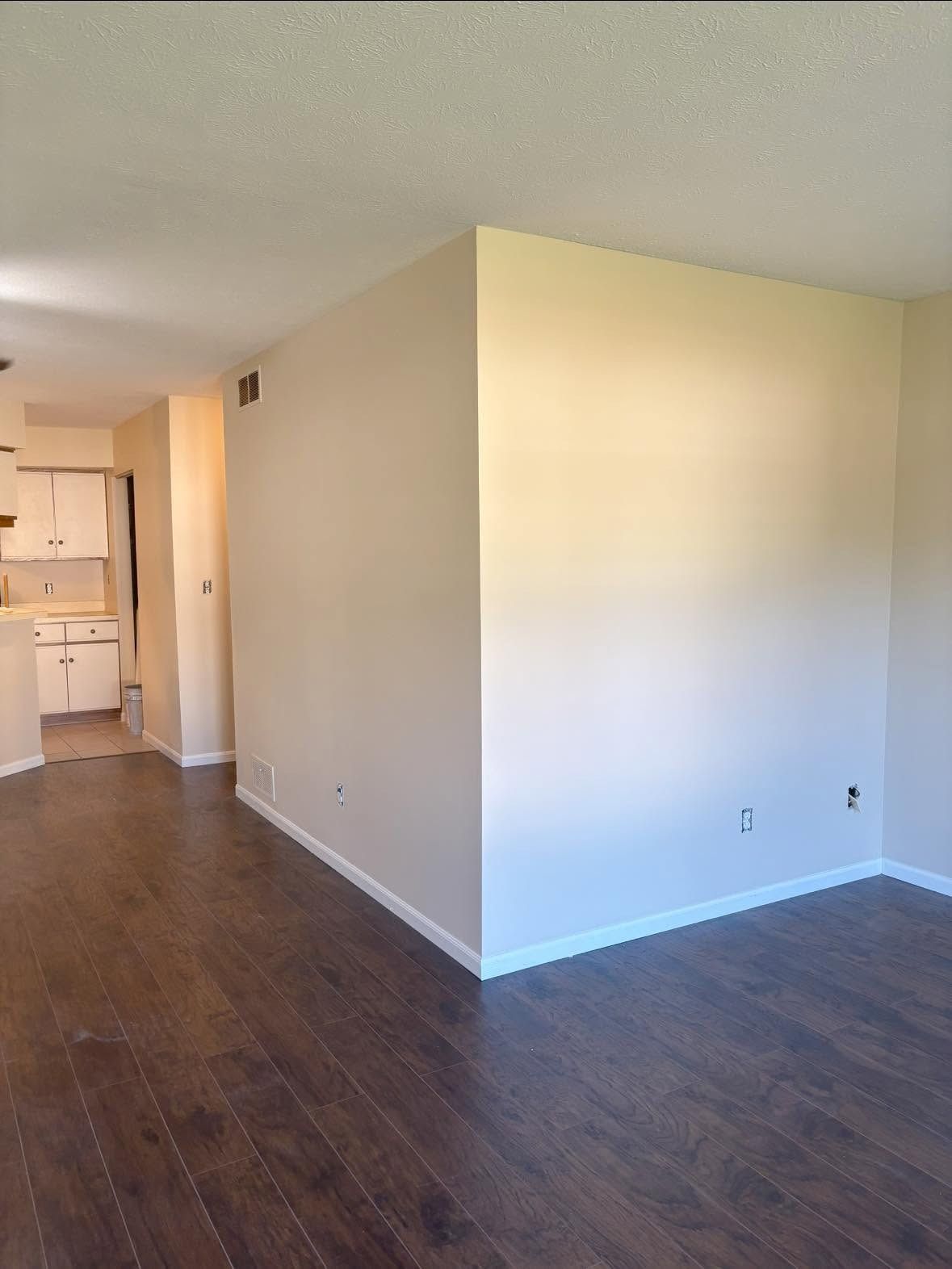 Empty apartment living room with dark wood floors, white walls, and kitchen visible in the background