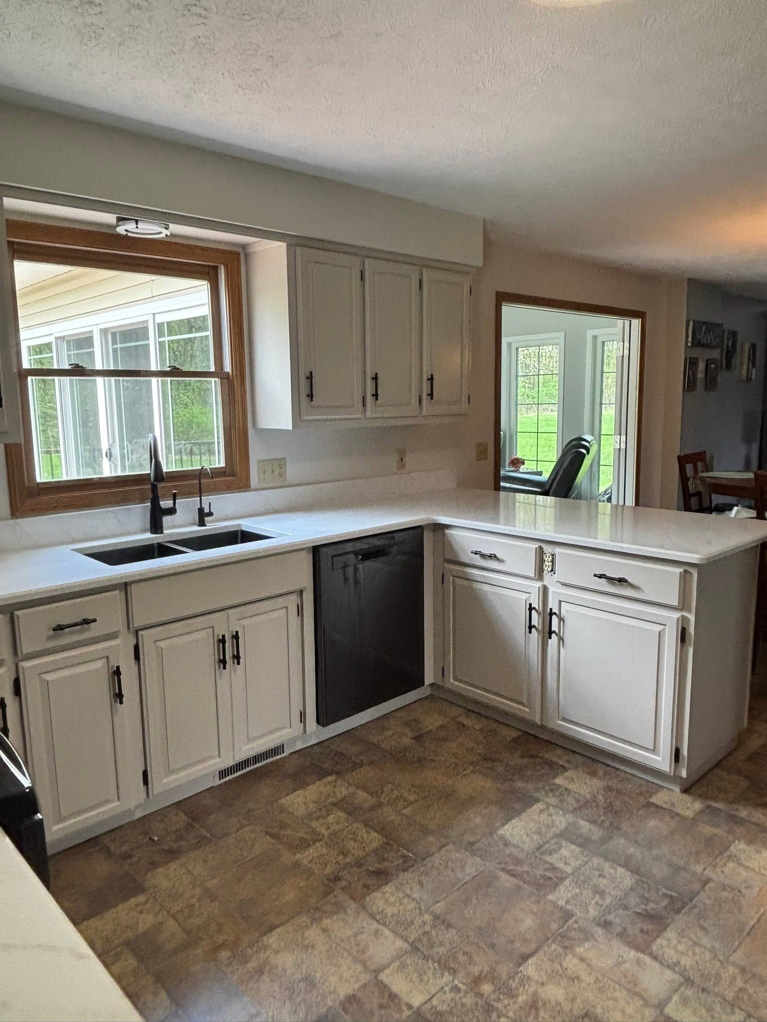 Empty kitchen with white cabinets, farmhouse sink, dark dishwasher, and wood-trimmed windows.