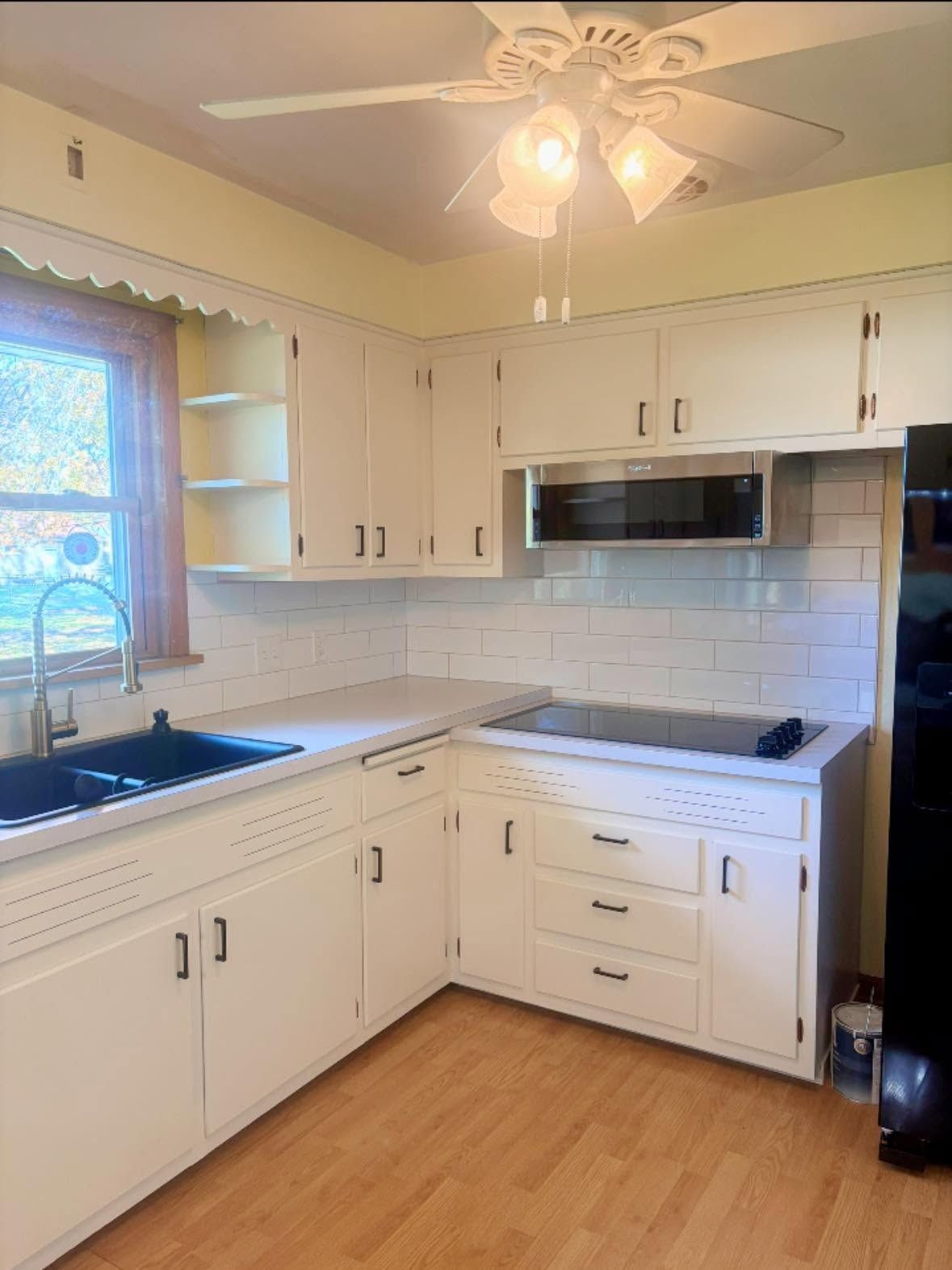 Bright kitchen with white cabinets, black countertops, tile backsplash, and wood floor under a ceiling fan.